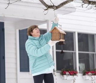 A woman wearing a winter coat hangs a bird house on her front porch