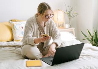 A woman sitting in bed looking at her phone with a laptop open in front of her