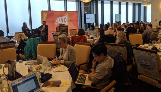 dozens of people seated at circular tables listening to a presentation