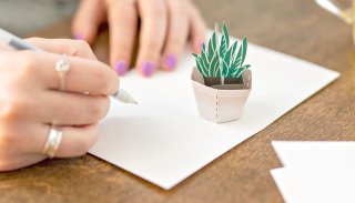 A woman writes out a note card with a paper, foldable succulent inside