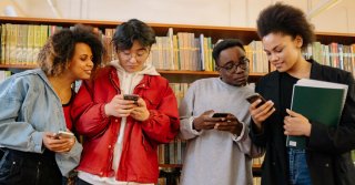 A group of prospective college students evaluating colleges with the help of AI on their mobile devices in front of a library bookshelf